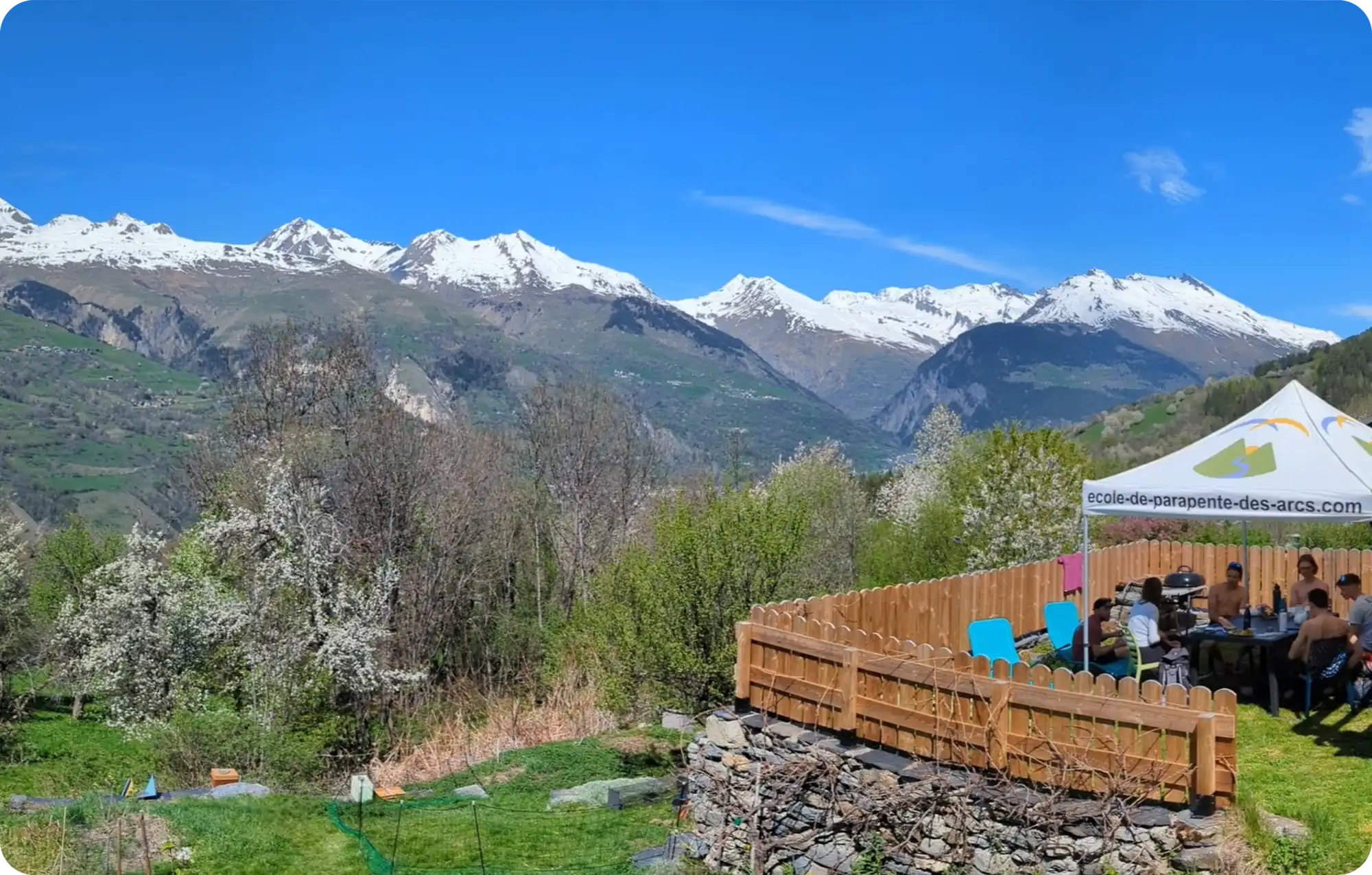 Terrasse du Gîte de l'Abeille à Montvenix avec vue panoramique sur les sommets enneigés des Alpes, jardin fleuri et groupe attablé sous un barnum de l'école de parapente des Arcs