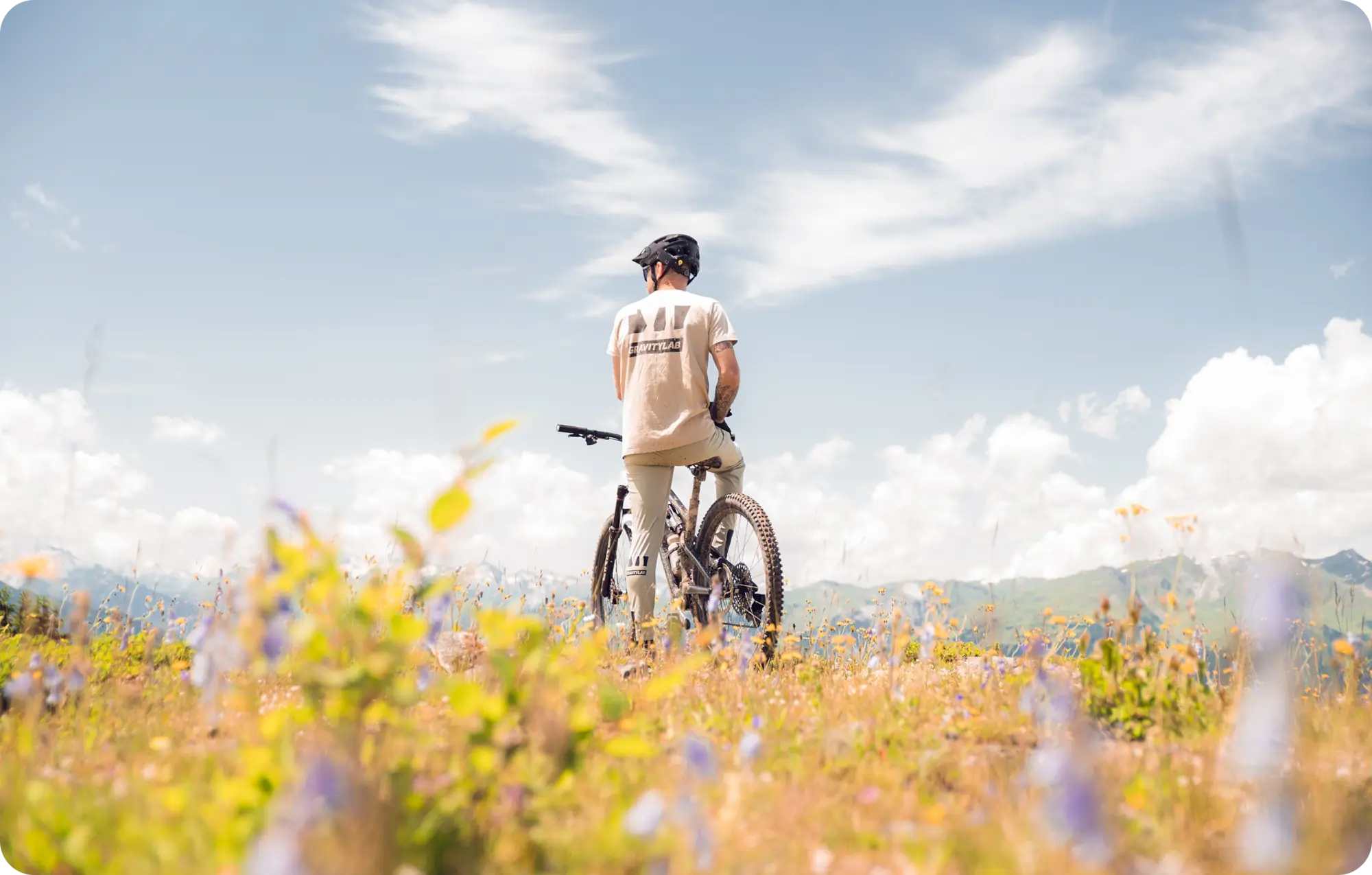 Cycliste de dos portant un t-shirt Gravitylab, posé sur son VTT au milieu d'une prairie alpine fleurie, face à un panorama de montagnes sous un ciel nuageux